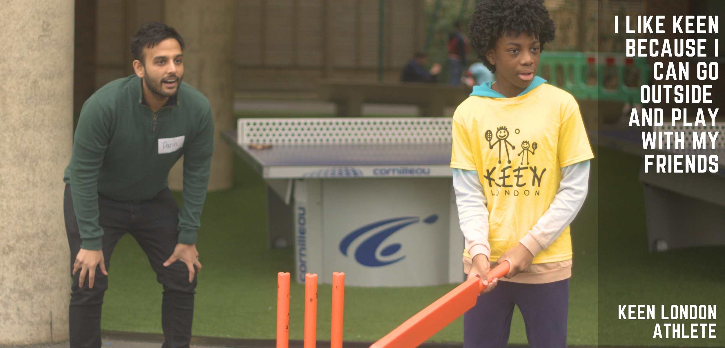 A determined KEEN London athlete, wearing a KEEN T-shirt, grips a cricket bat, with his dedicated volunteer coach standing beside him, eagerly anticipating the incoming ball.